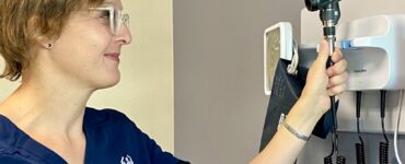 Dr. Verena Samara, a neurologist, wearing a navy blue medical uniform with her name and title embroidered, reaching to adjust an otoscope on a wall-mounted medical device in a clinic room.