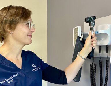 Dr. Verena Samara, a neurologist, wearing a navy blue medical uniform with her name and title embroidered, reaching to adjust an otoscope on a wall-mounted medical device in a clinic room.