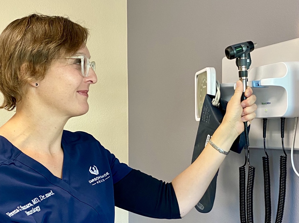 Dr. Verena Samara, a neurologist, wearing a navy blue medical uniform with her name and title embroidered, reaching to adjust an otoscope on a wall-mounted medical device in a clinic room.