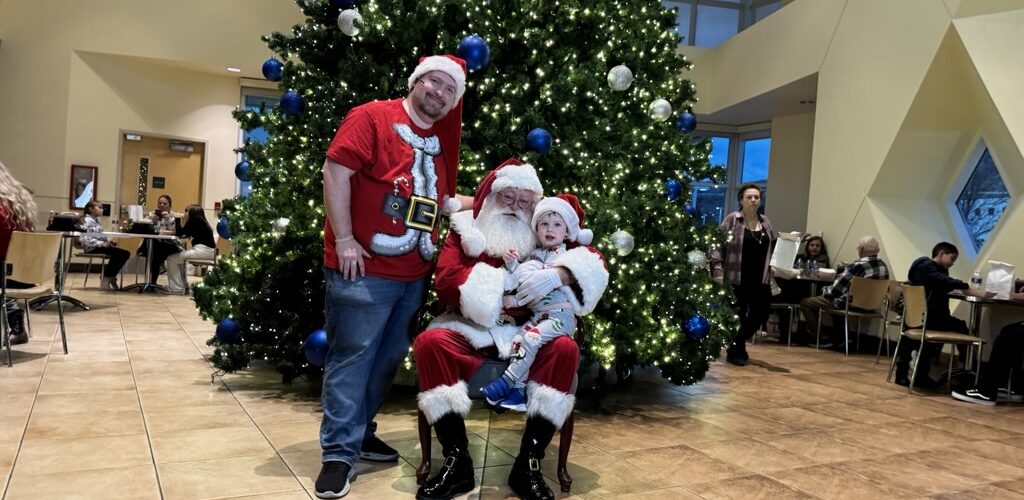Child sitting with Santa Claus in front of a large decorated Christmas tree during a holiday celebration at Carson Tahoe Health.