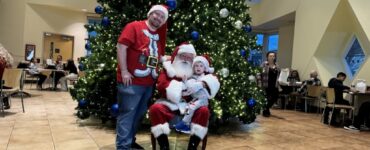 Child sitting with Santa Claus in front of a large decorated Christmas tree during a holiday celebration at Carson Tahoe Health.
