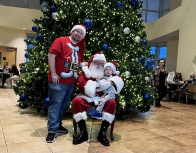 Child sitting with Santa Claus in front of a large decorated Christmas tree during a holiday celebration at Carson Tahoe Health.