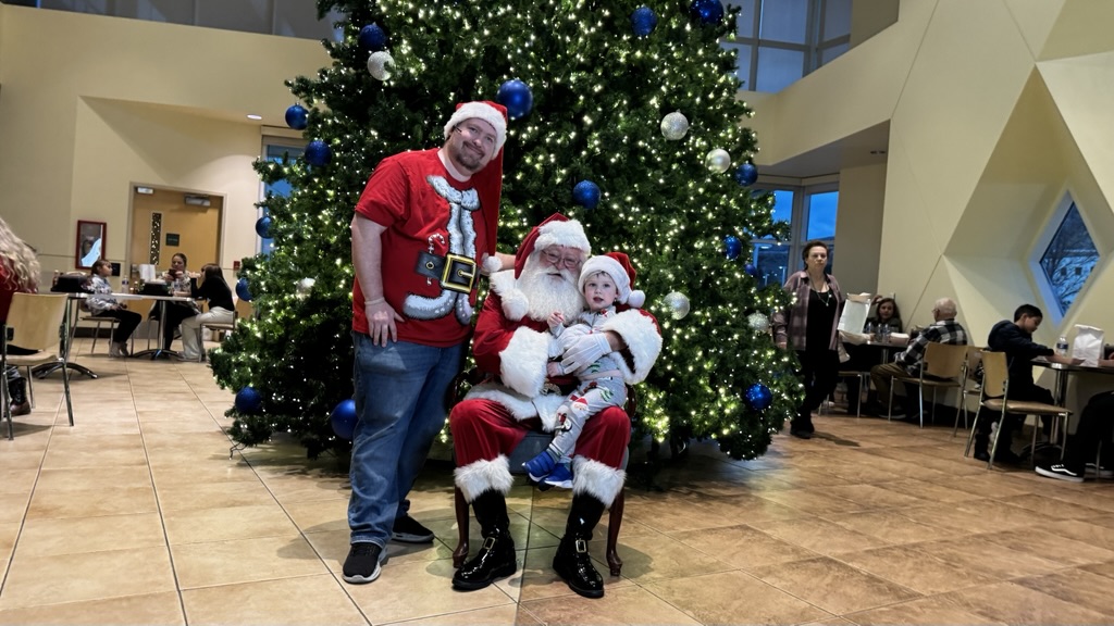Child sitting with Santa Claus in front of a large decorated Christmas tree during a holiday celebration at Carson Tahoe Health.