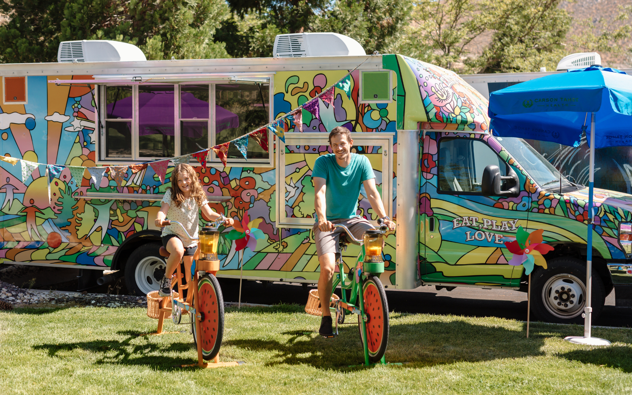 An adult and child riding stationary bikes in front of Carson Tahoe Health’s colorful Vehicles of Change mobile program van.