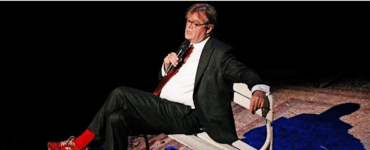 Storyteller performing on stage while seated on a bench, holding a microphone under stage lighting.