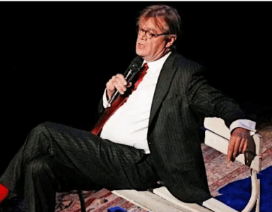 Storyteller performing on stage while seated on a bench, holding a microphone under stage lighting.