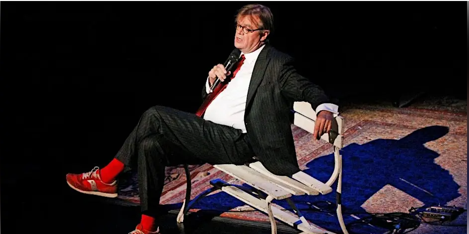 Storyteller performing on stage while seated on a bench, holding a microphone under stage lighting.