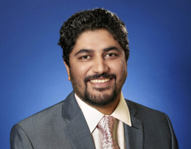 Cardiologist wearing a suit and tie, smiling against a blue studio background