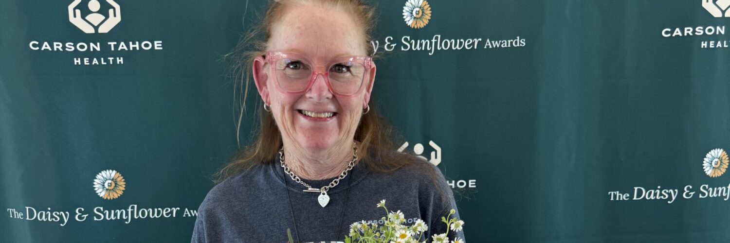 DAISY Award honoree holding two bouquets of flowers while standing in front of a Carson Tahoe Health DAISY & Sunflower Awards backdrop.