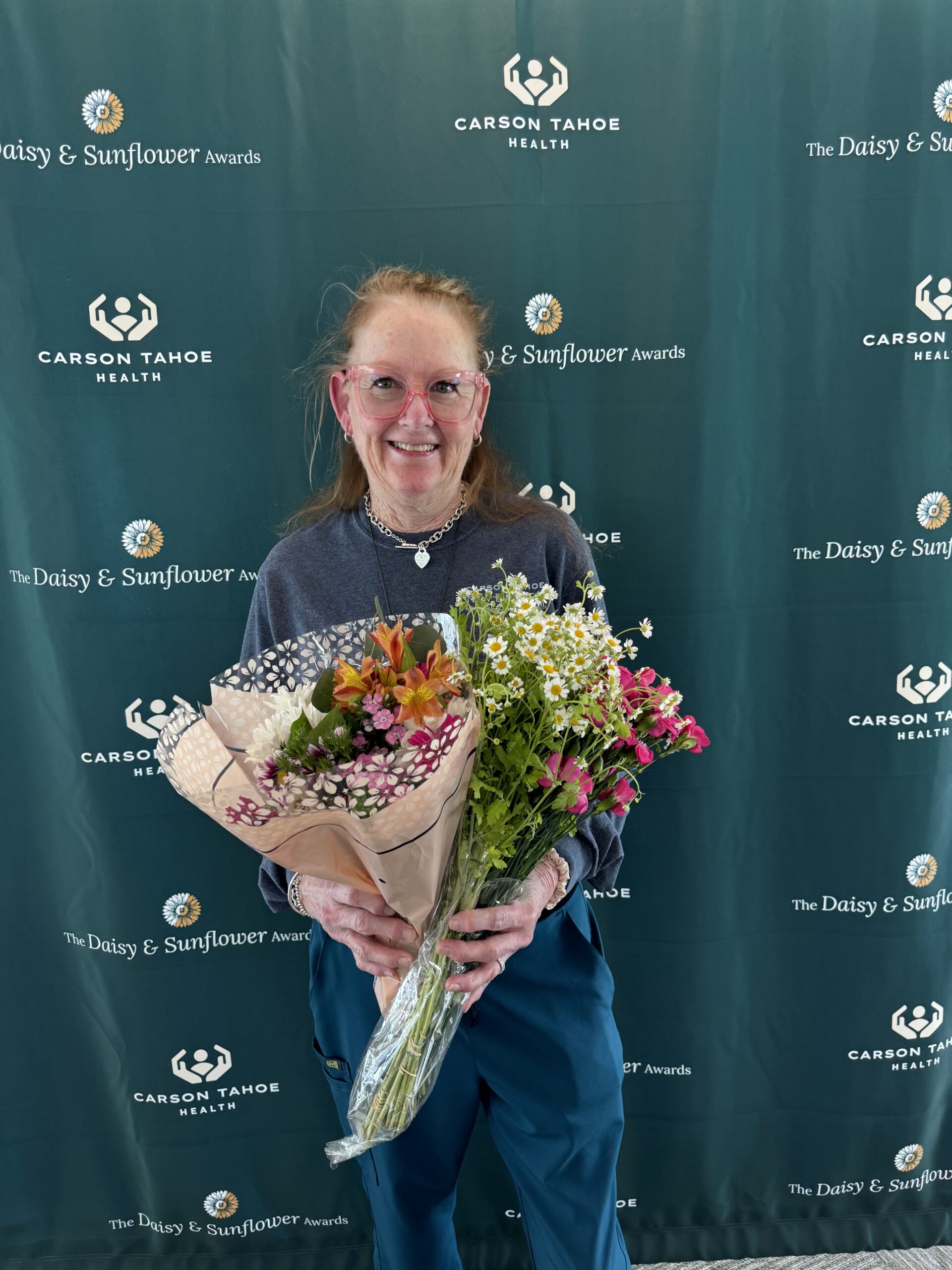 DAISY Award honoree holding two bouquets of flowers while standing in front of a Carson Tahoe Health DAISY & Sunflower Awards backdrop.