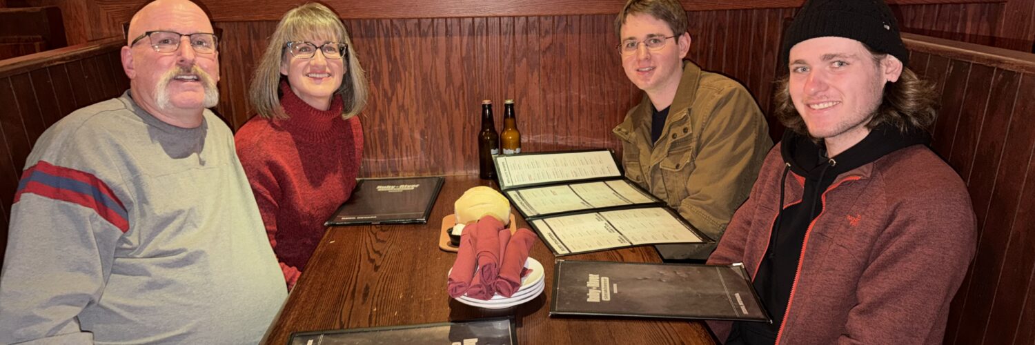 Four people seated together at a restaurant booth, smiling at the camera with menus on the table.
