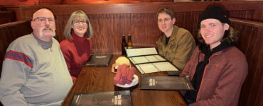 Four people seated together at a restaurant booth, smiling at the camera with menus on the table.