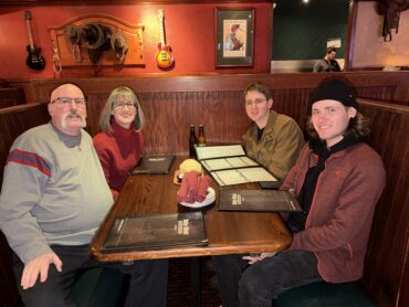 Four people seated together at a restaurant booth, smiling at the camera with menus on the table.
