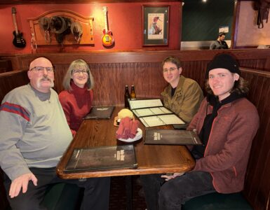 Four people seated together at a restaurant booth, smiling at the camera with menus on the table.