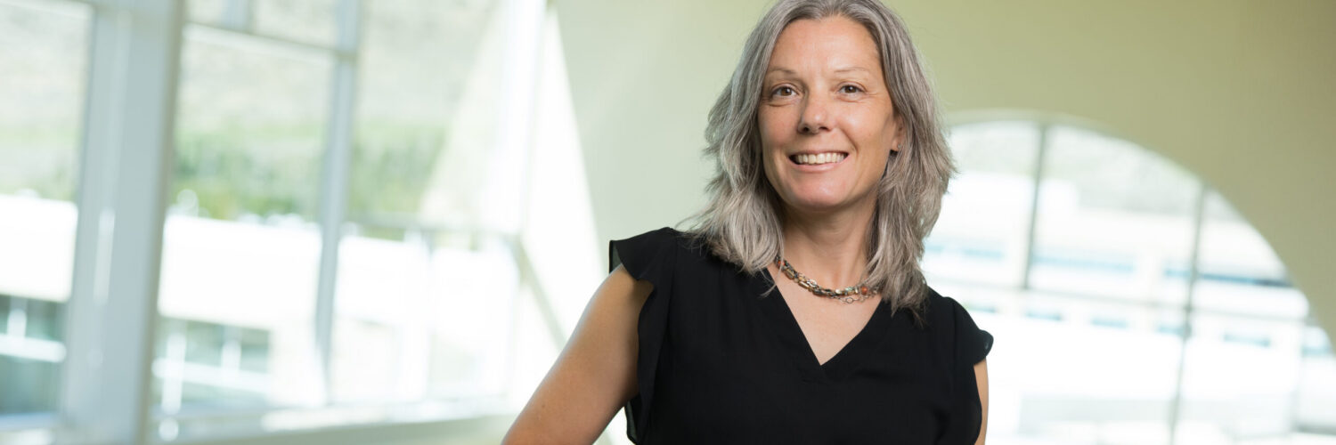 Registered dietitian standing indoors at a hospital facility, smiling and leaning on a railing with large windows in the background