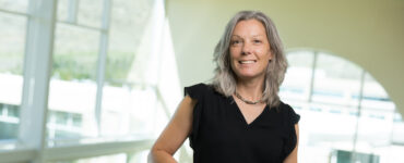 Registered dietitian standing indoors at a hospital facility, smiling and leaning on a railing with large windows in the background