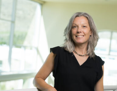 Registered dietitian standing indoors at a hospital facility, smiling and leaning on a railing with large windows in the background