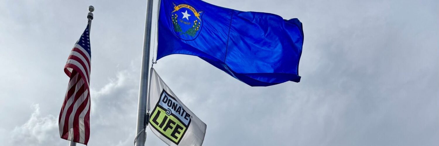 Donate Life flag and U.S. flag raised on flagpoles outside Carson Tahoe Regional Medical Center building under a cloudy sky.