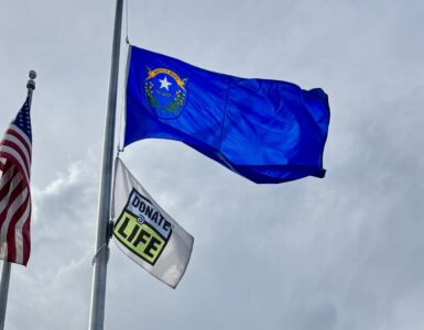 Donate Life flag and U.S. flag raised on flagpoles outside Carson Tahoe Regional Medical Center building under a cloudy sky.