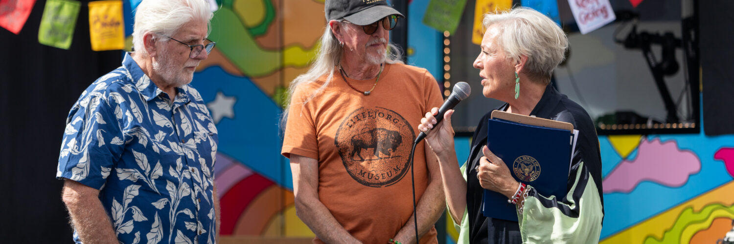 A woman speaks into a microphone while presenting a recognition to two men in front of Carson Tahoe Health's colorful SOUND IDEAS outreach vehicle during the Dave Mason legacy dedication ceremony.