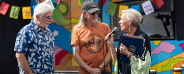 A woman speaks into a microphone while presenting a recognition to two men in front of Carson Tahoe Health's colorful SOUND IDEAS outreach vehicle during the Dave Mason legacy dedication ceremony.