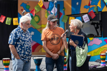 A woman speaks into a microphone while presenting a recognition to two men in front of Carson Tahoe Health's colorful SOUND IDEAS outreach vehicle during the Dave Mason legacy dedication ceremony.