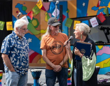 A woman speaks into a microphone while presenting a recognition to two men in front of Carson Tahoe Health's colorful SOUND IDEAS outreach vehicle during the Dave Mason legacy dedication ceremony.